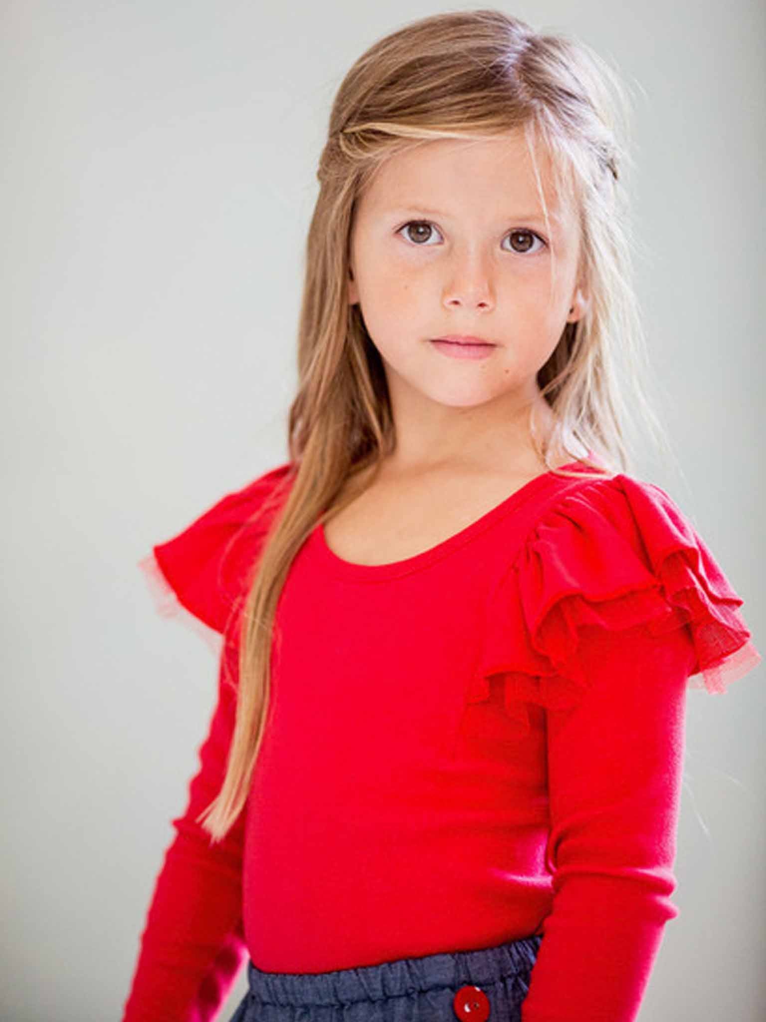 A young girl with long hair wears the Essentials Sweetheart Red Tee - L/S with net flutter sleeves and a dark skirt, looking directly at the camera.