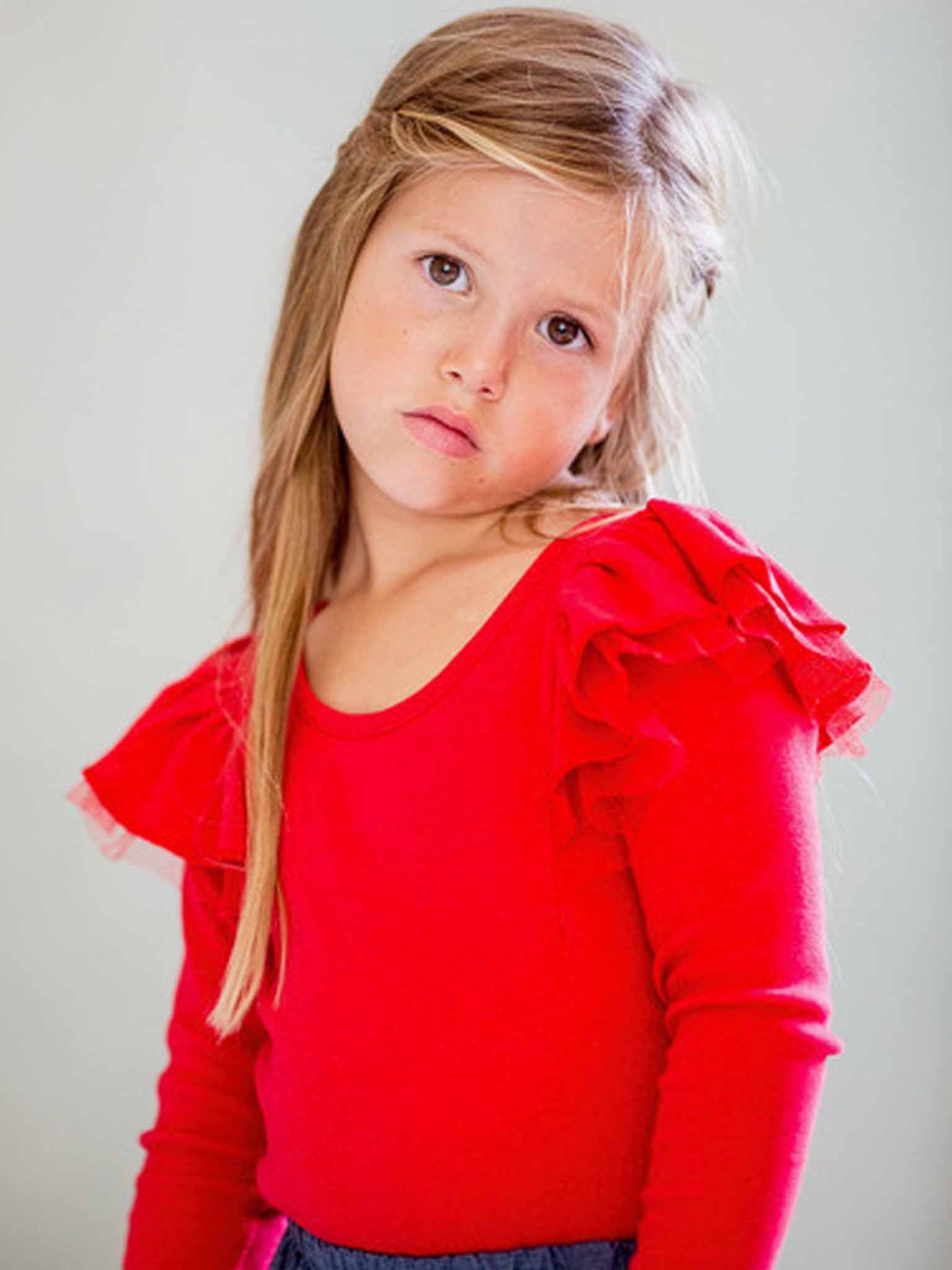 A young girl with long hair, dressed in an Essentials Sweetheart Red Tee - L/S, poses gracefully against a neutral background.