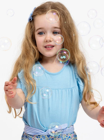 A young girl with long, wavy blonde hair and a Gathered Aqua Blue Tee from Essentials, designed in an easy-wear 100% cotton style, is reaching out towards floating soap bubbles.