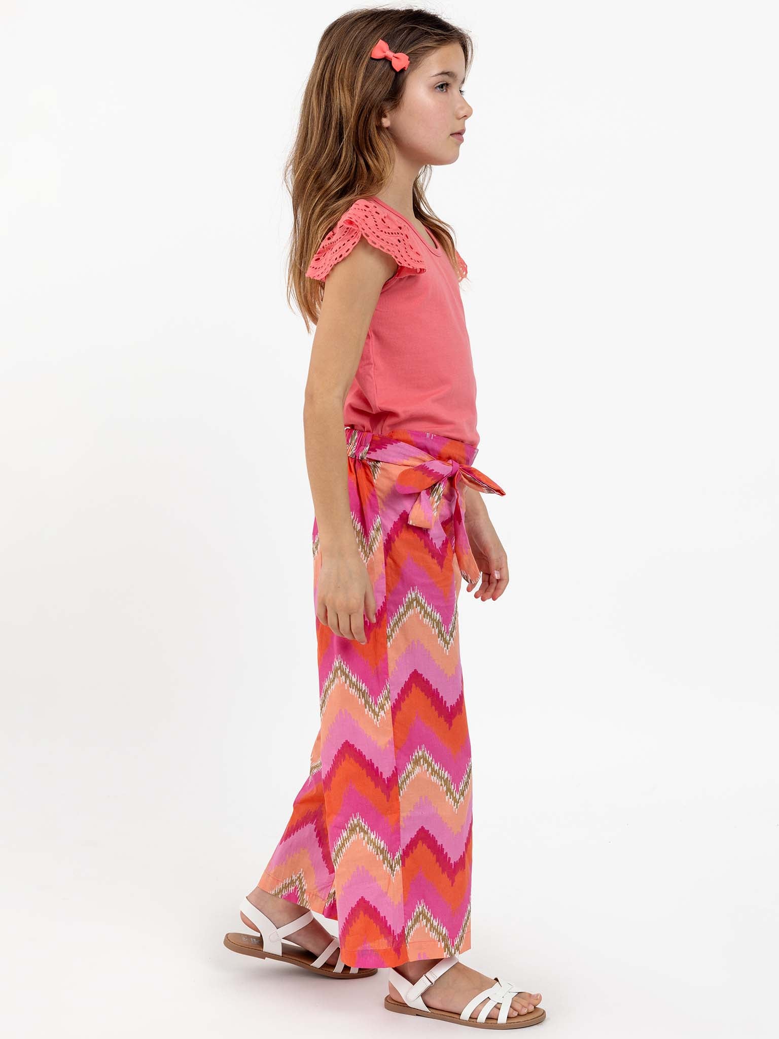 A young girl with long hair in a pink top and Golden Ikat Trousers, which feature a comfy, elastic waistband and colorful zigzag patterns, stands facing sideways. She wears white sandals and has a pink hair clip.