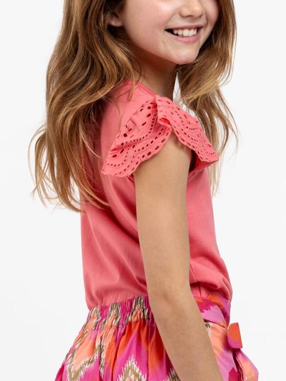 A young girl smiles while wearing an Essentials Lace Sleeve Primrose Pink Tee made of 100% cotton and a colorful patterned skirt, photographed from the side.