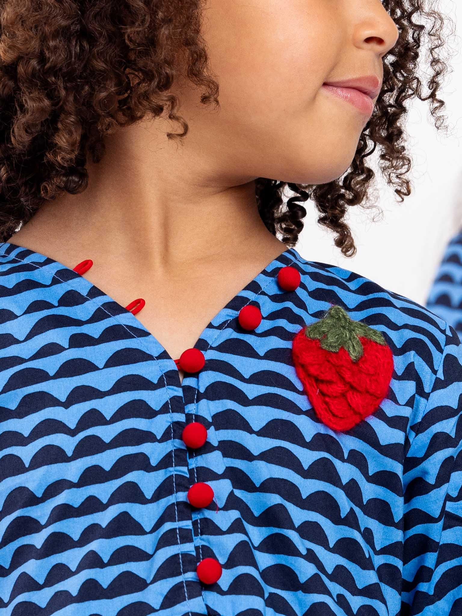 Close-up of a woman in a Wiggly Ocean Blue Adriana dress with red buttons, featuring a large red strawberry brooch on the collar.