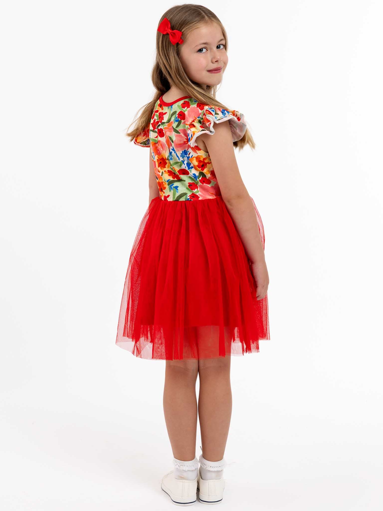 A young girl stands against a plain background, dressed in the vibrant Wild Roses Ariel Dress. This charming party piece features a colorful Wild Roses print top paired with a red tulle skirt. Her stretch jersey bodice offers comfort, while white shoes and a red hair bow complete the delightful look from Wild Roses.