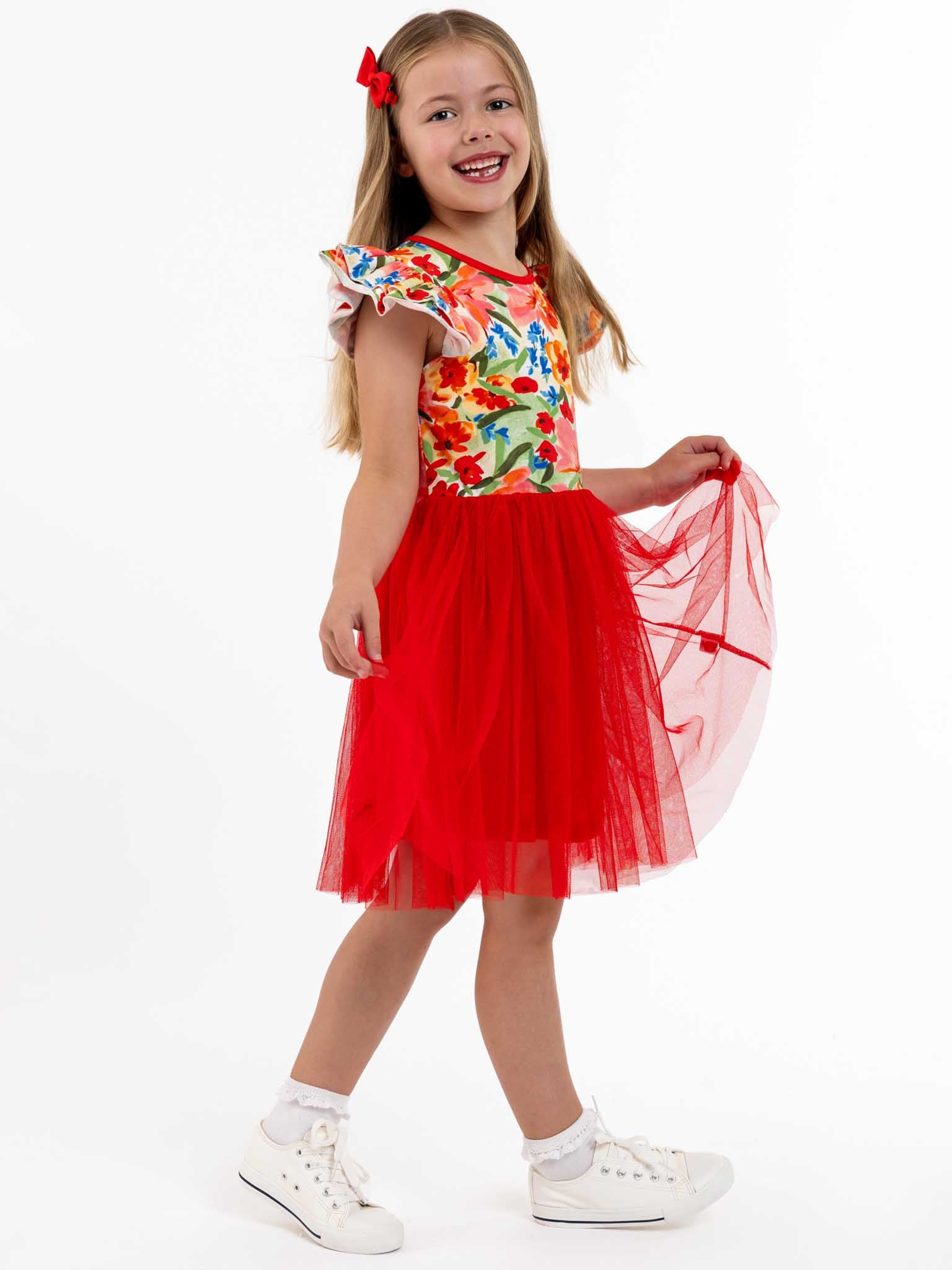 A young girl in a vibrant dress featuring a red tulle skirt cheerfully poses against a white background, wearing the Wild Roses Chloe Ponytail Hat with a wide brim and contrast fabric lining, paired with white sneakers.