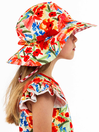 Child in profile wearing a Wild Roses Chloe Ponytail Hat adorned with a colorful floral pattern, paired with a dress showcasing red, blue, and orange designs.