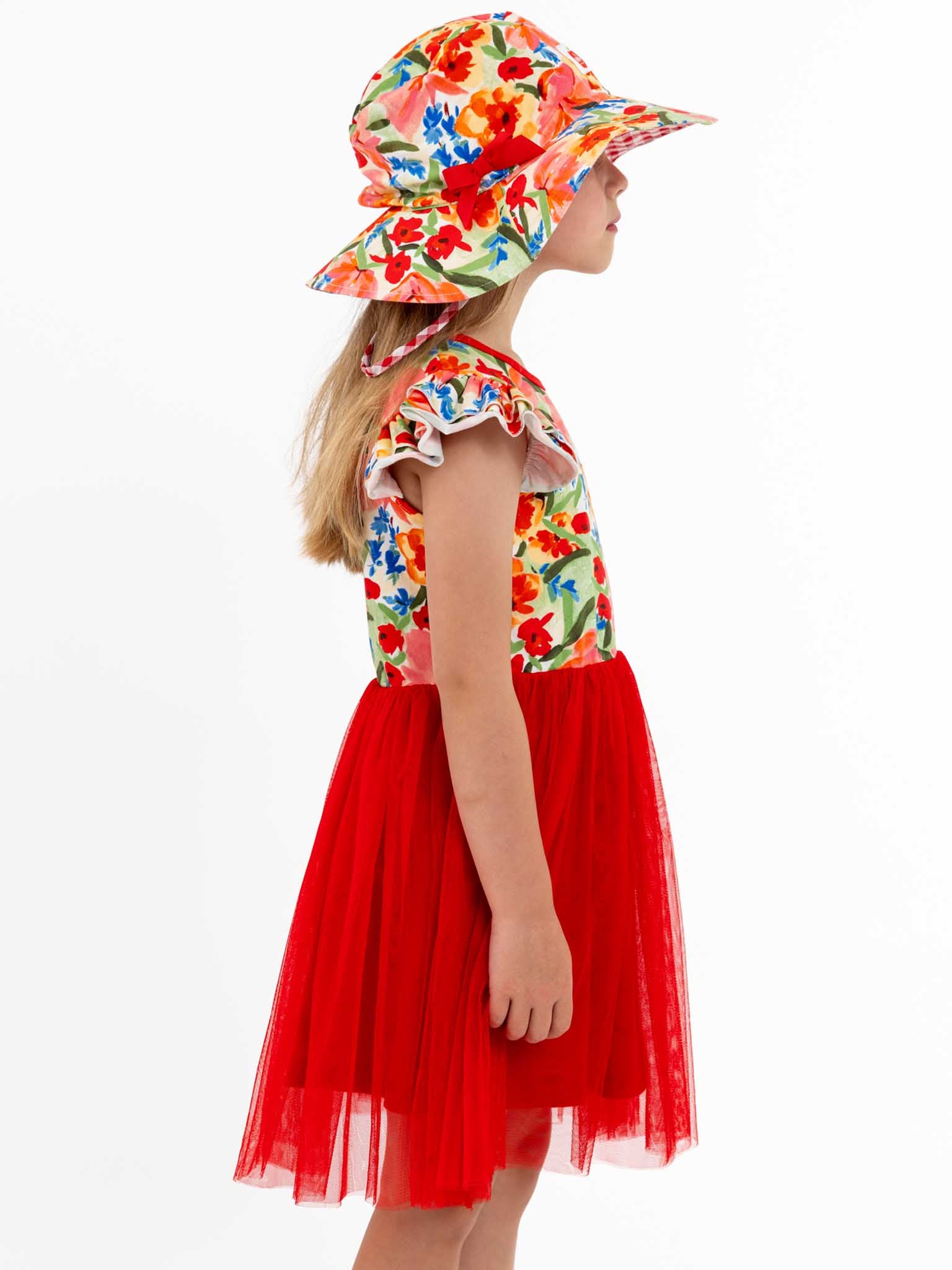 A girl in a red skirt wearing a Wild Roses wide-brim hat with the Chloe ponytail style, featuring a floral pattern, is facing sideways.