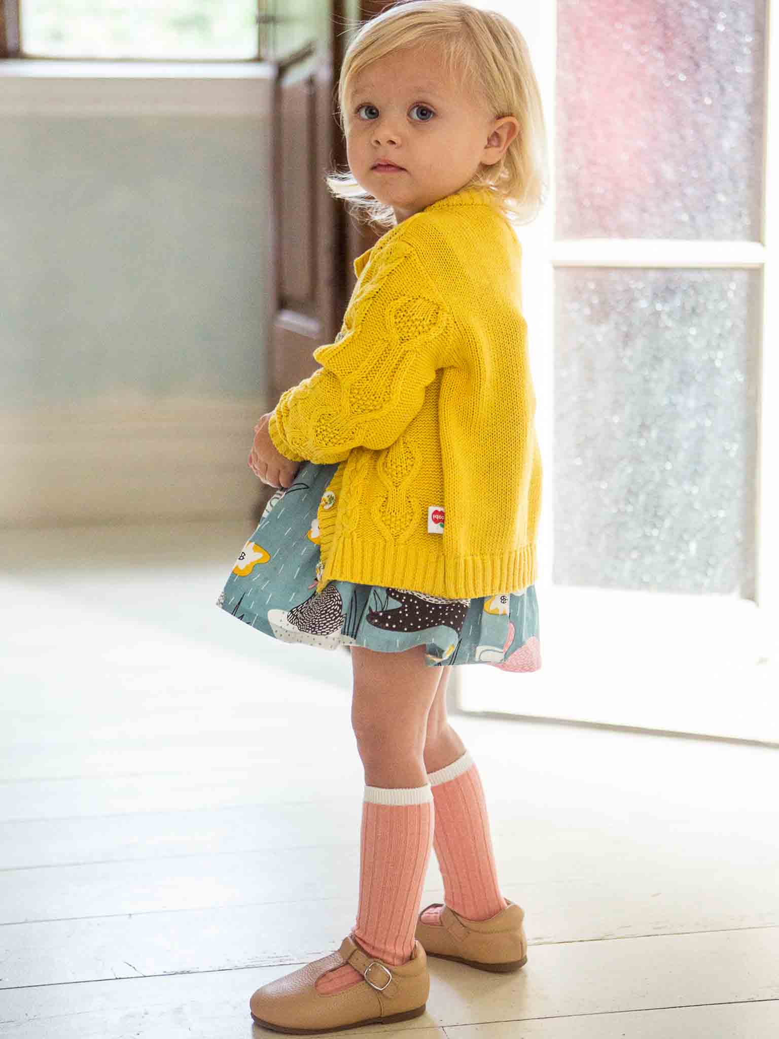 Toddler in a yellow cable knit Knitwear Willow Sunshine Cotton Cardigan and patterned skirt standing indoors.