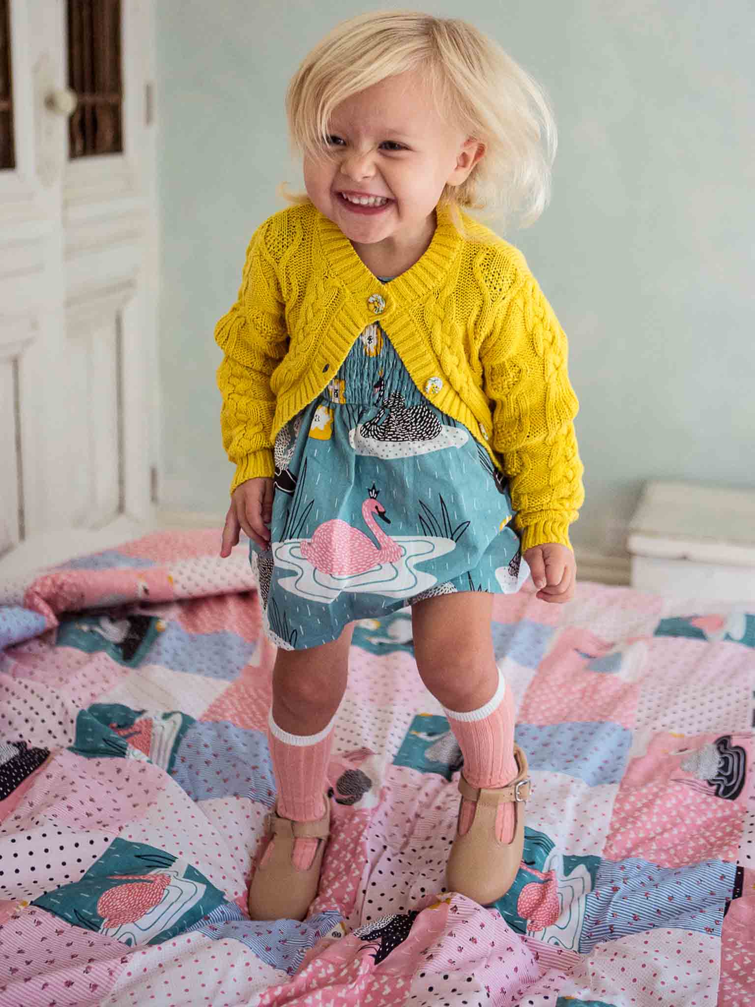 Young child smiling in a cable-knit Willow Sunshine Cotton Cardigan and patterned dress standing on a bed with a colorful quilt.