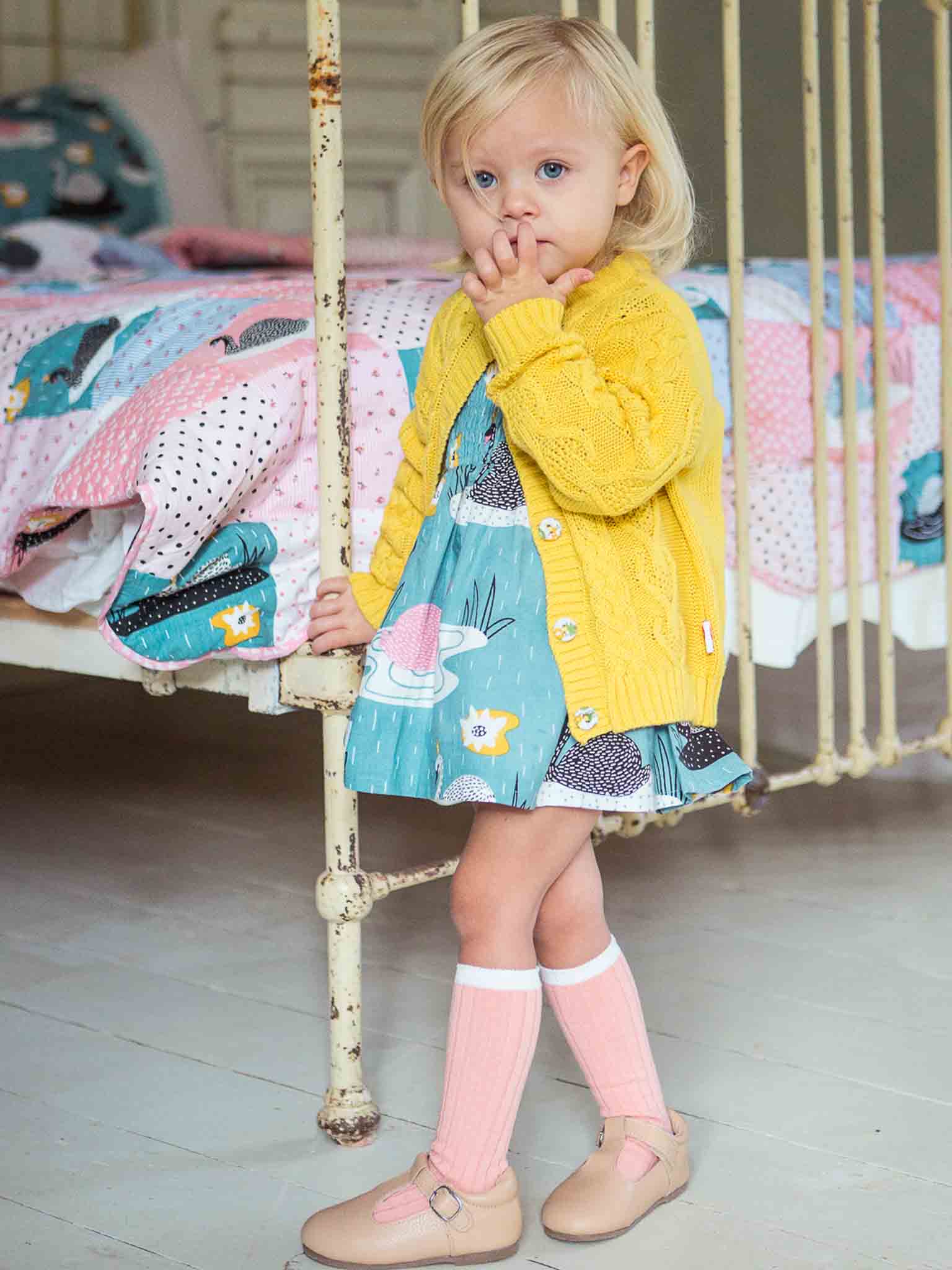 A young child in a Willow Sunshine Cotton Cardigan by Knitwear and printed dress, standing by a white crib with a thoughtful expression.