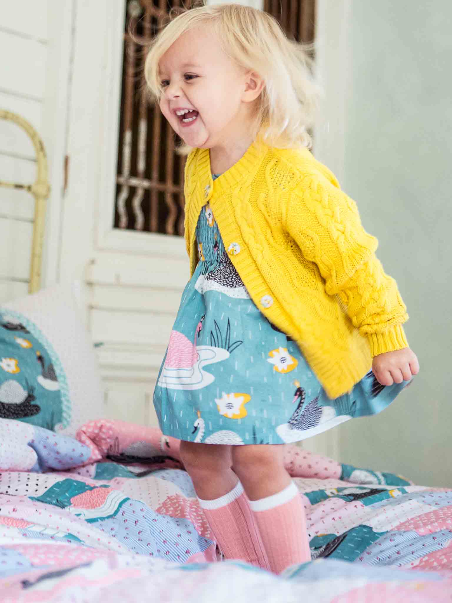 A young child in a high-quality 100% cotton cable knit Willow Sunshine Cotton Cardigan and patterned dress laughing while standing on a bed with a colorful quilt.