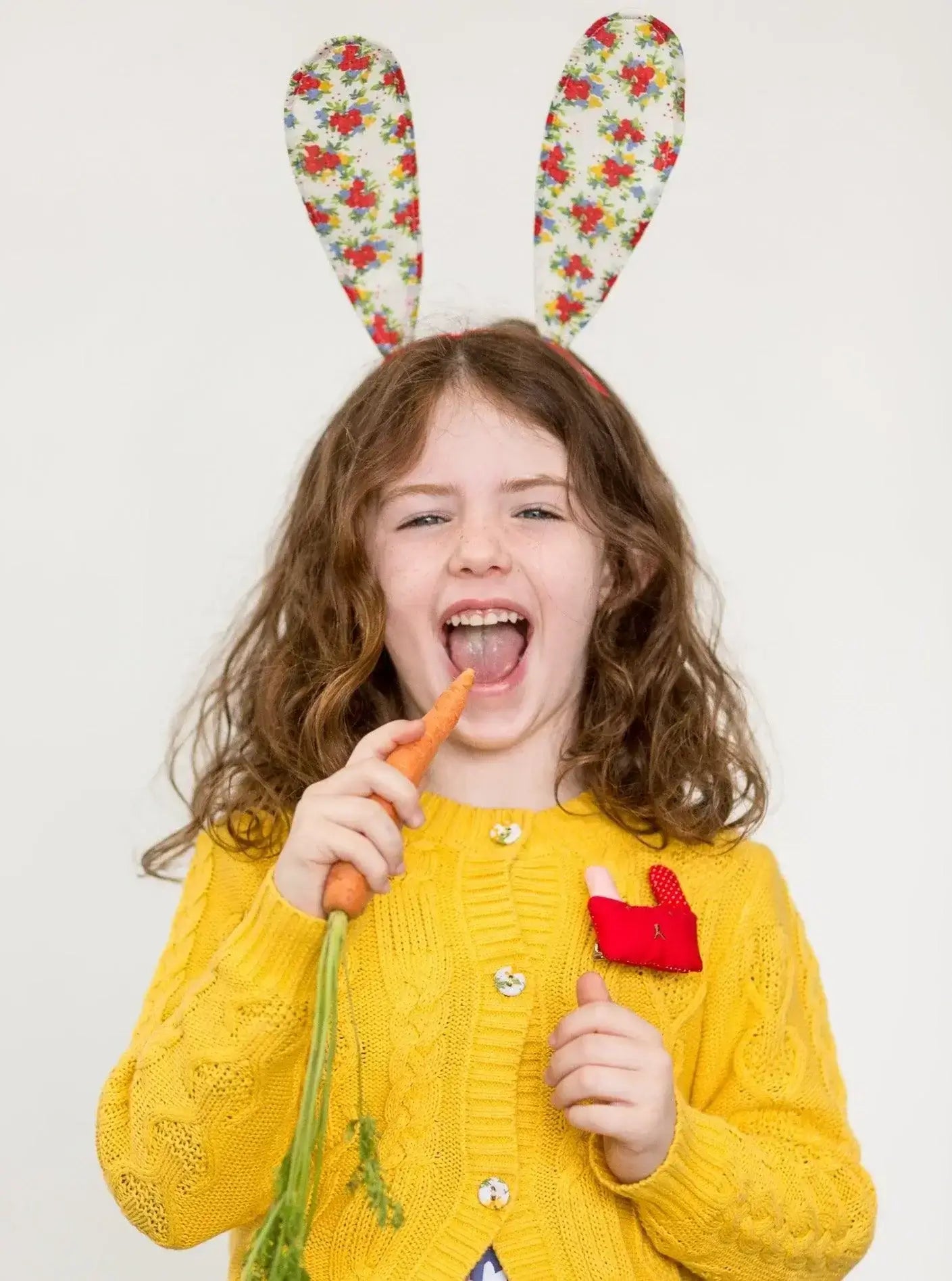 A little girl wearing the Willow Sunshine Cotton Cardigan from Knitwear, cotton cable knit bunny ears and holding a carrot, featuring fabric covered buttons.