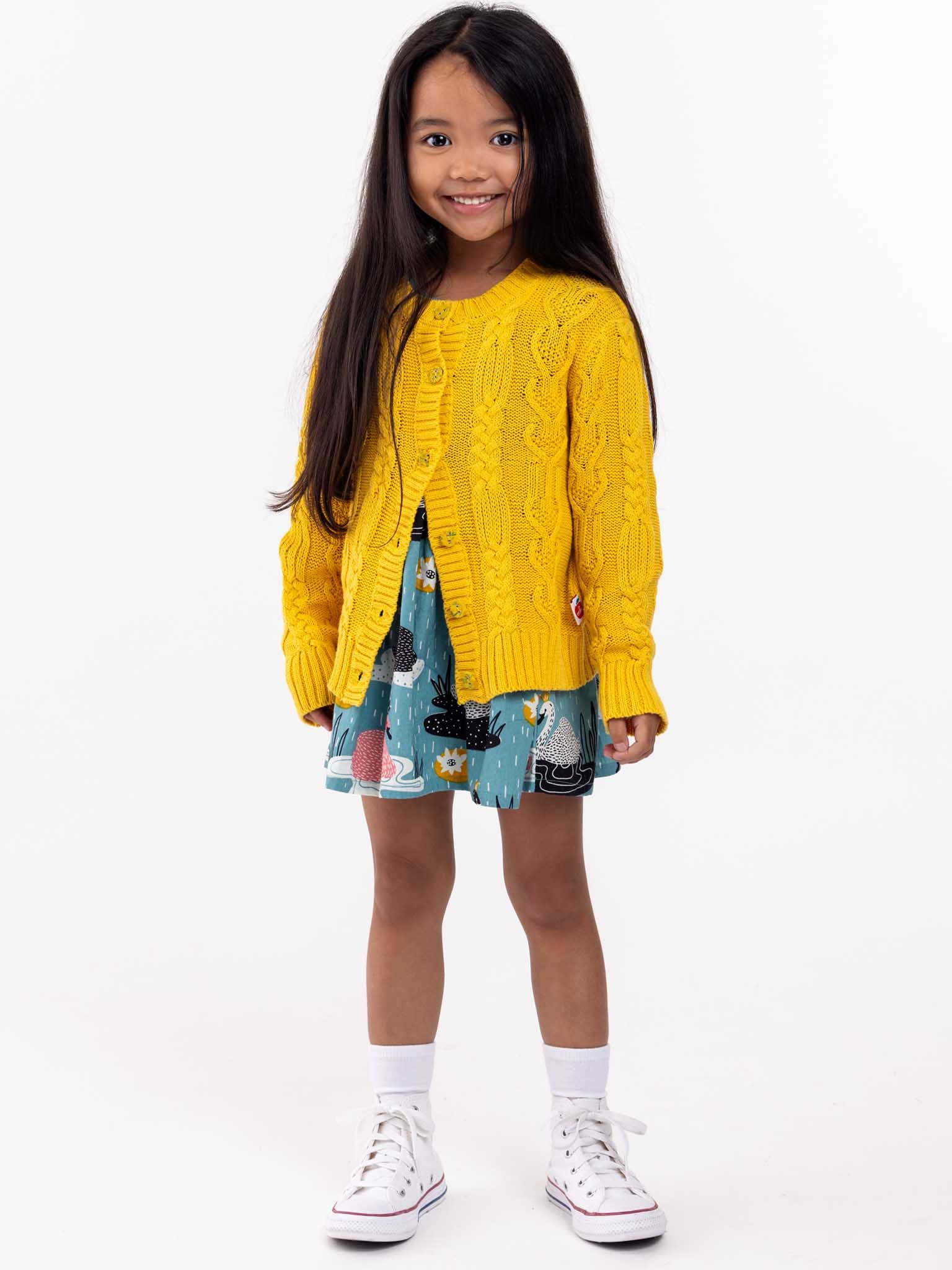 Young girl smiling, wearing a bright yellow Knitwear Willow Sunshine Cotton Cardigan, patterned dress, and white sneakers, standing against a white background.