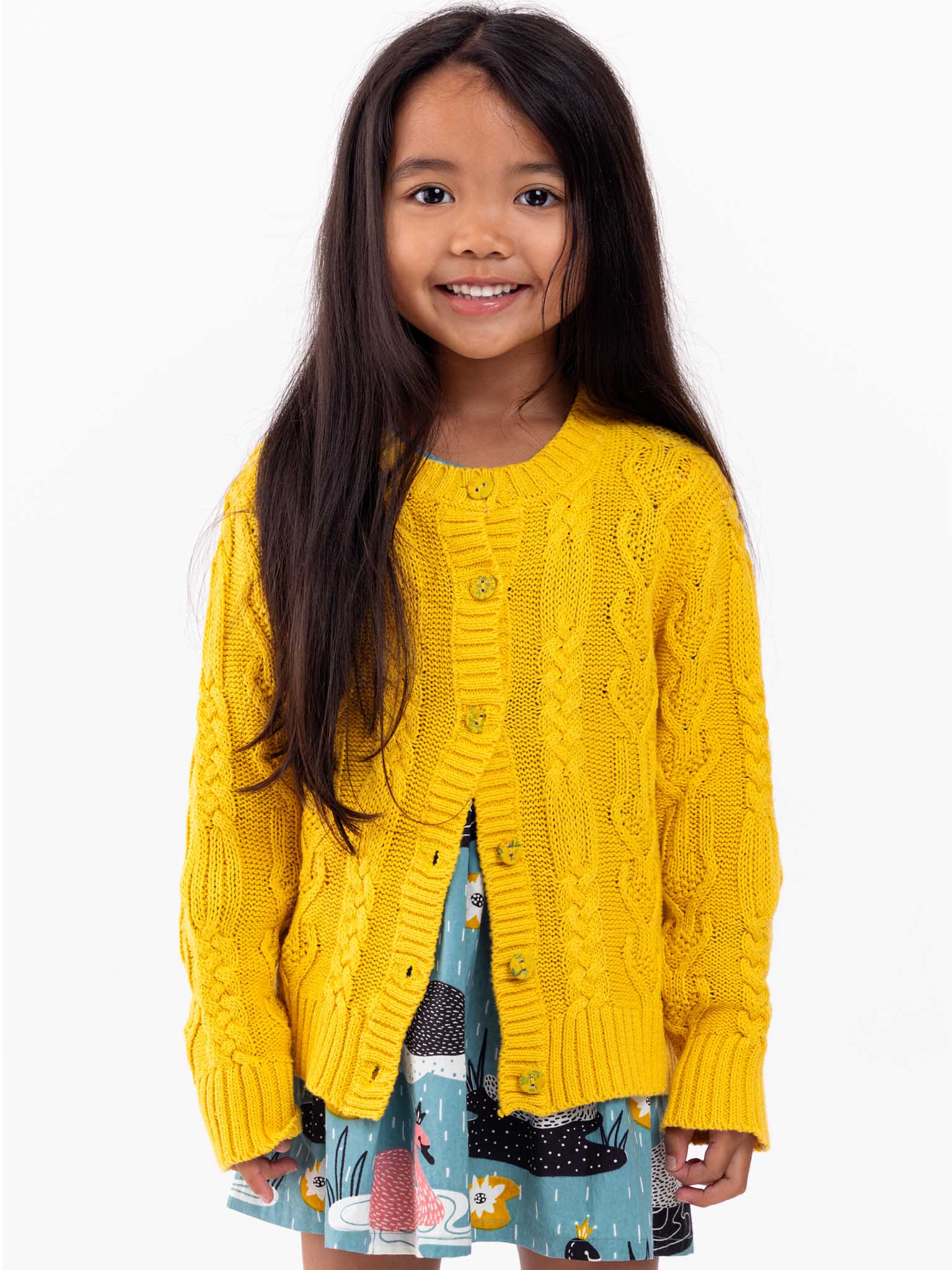 Young girl with long brown hair, wearing a yellow cable knit Willow Sunshine Cotton Cardigan and a colorful skirt, smiling at the camera on a white background.