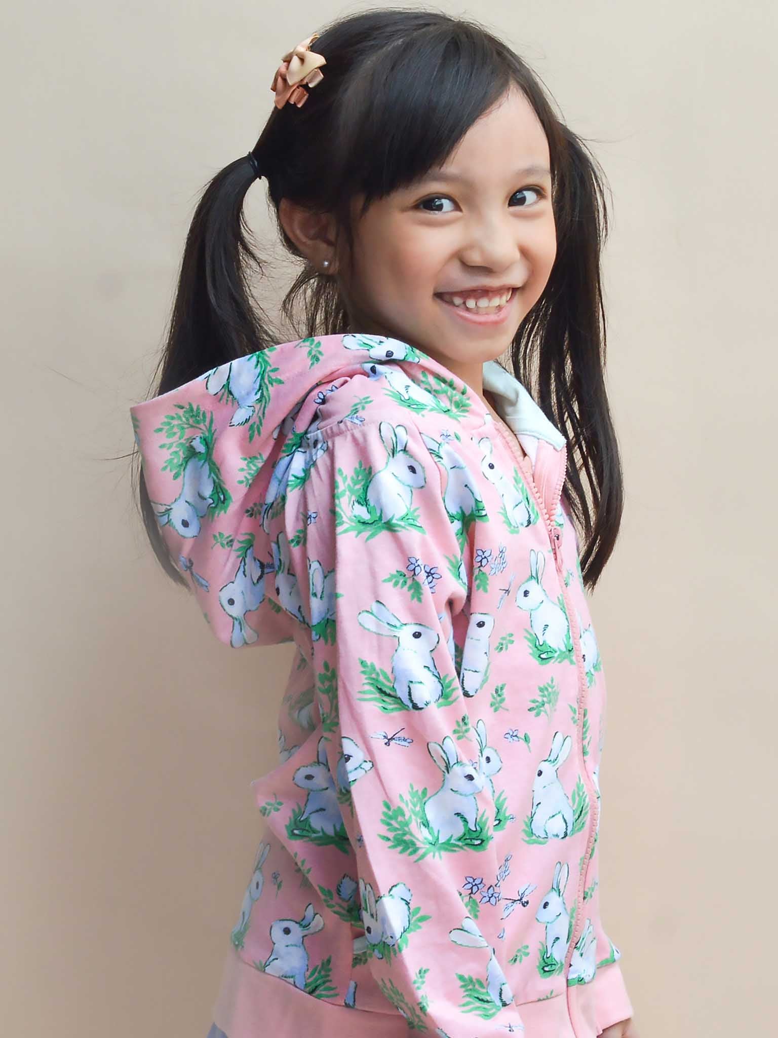 A girl with long black pigtails adorably smiles in the Pink Bun-Buns Zadie Jacket, featuring white rabbit prints, while standing sideways against a plain background.