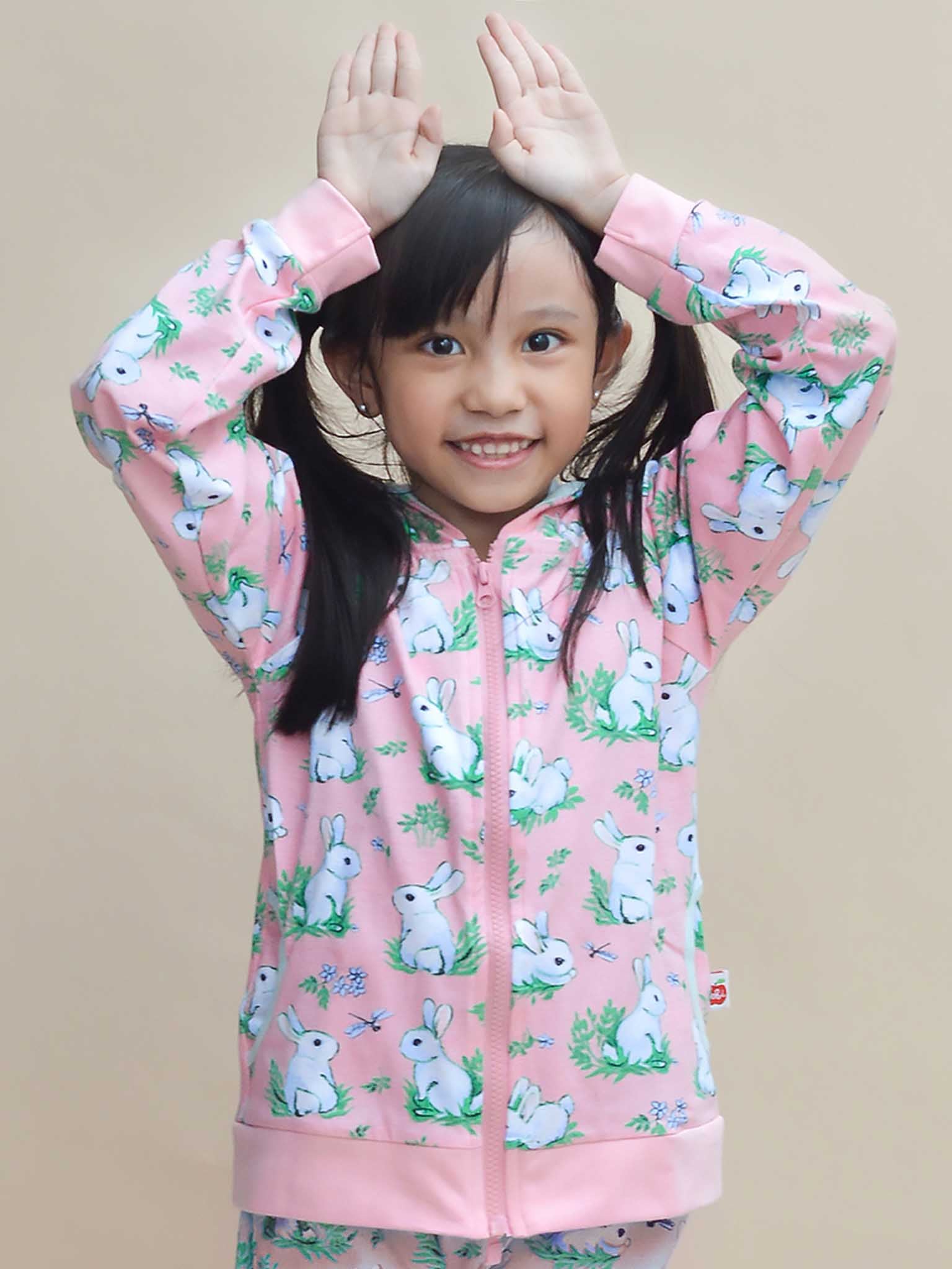 A child grins, hands as bunny ears, wearing the Pink Bun-Buns Zadie Jacket from Bun-Buns, adorned with a rabbit pattern. Their pink sweatshirt complements the soft jersey pants, crafting a charming outfit.