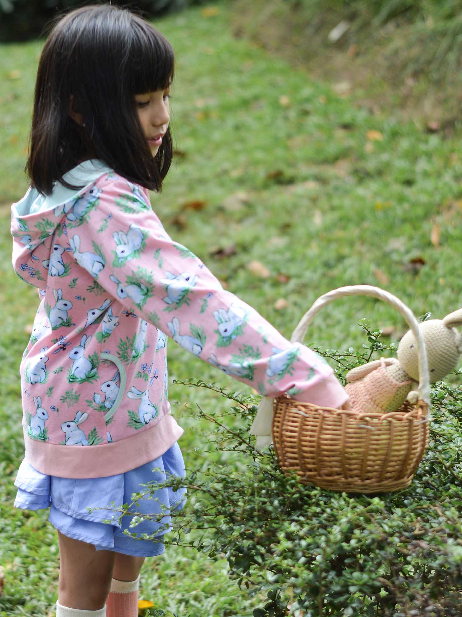 A child wearing a Bun-Buns Pink Bun-Buns Zadie Jacket and matching soft jersey pants places a stuffed animal in a wicker basket on the grass.