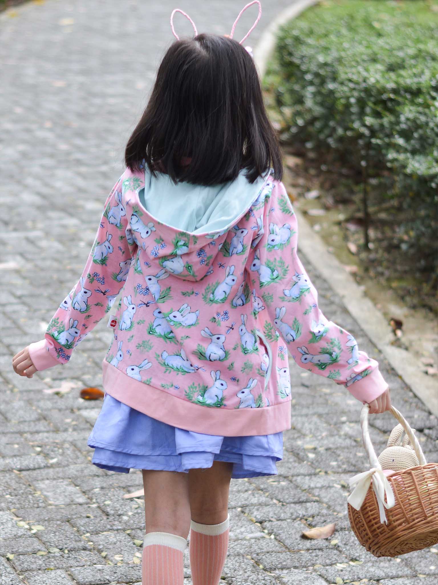 A child in a Bun-Buns Pink Bun-Buns Zadie Jacket and blue skirt walks on a stone path, holding a wicker basket, perfectly complementing her adorable snuggly jacket.