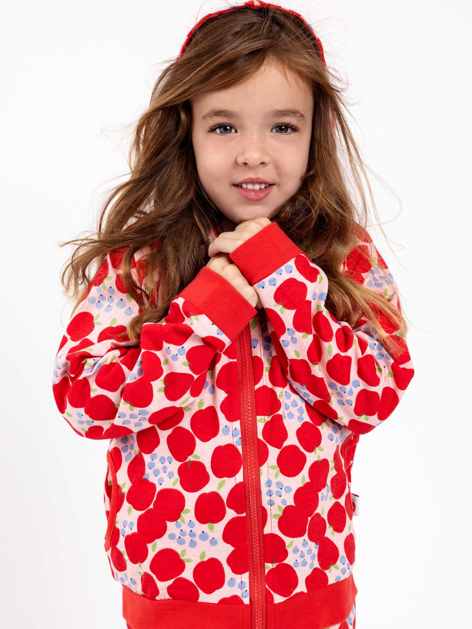 A child with clasped hands and long brown hair wears the Apple Zadie Jacket from Sweet Apple, featuring a red and white polka dot hood.